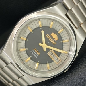 May include: A silver-tone stainless steel wristwatch with a dark gray circular dial, gold-tone hands, and gold-tone hour markers. The dial features the Orient logo, the text AAA Crystal 21 JEWELS, and a day-date window displaying TUE 7.