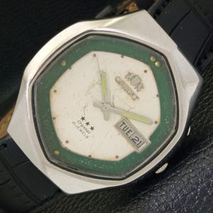 Peut inclure: Une montre-bracelet argentée avec un cadran vert et un bracelet en cuir noir. Le cadran de la montre a un fond blanc avec des aiguilles noires, une fenêtre de date qui affiche "TUE 21", et le texte "Orient Crystal 21 Jewels".