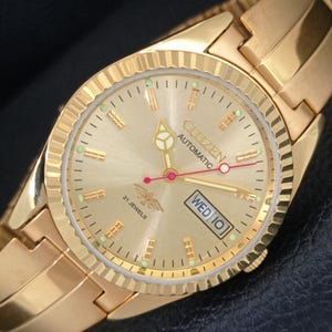 Op de afbeelding: Een goudkleurig Citizen automatisch polshorloge met een champagnekleurige wijzerplaat met zonnestraalpatroon, goudkleurige streepindexen, lichtgevende accenten en een dag-datumvenster op de drie-uurpositie. Het horloge heeft een geribbelde lunette.
