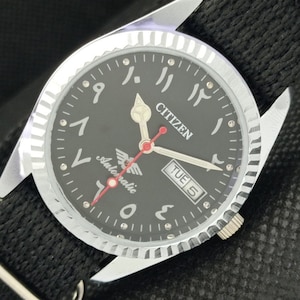Può includere: Un orologio automatico Citizen con quadrante nero, numeri arabi e visualizzazione del giorno della settimana. L'orologio ha una cassa argentata e un cinturino in nylon nero.