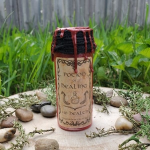 Puede incluir: Una pequeña botella de poción roja con una etiqueta que dice "Potion of Healing". La parte superior está sellada con hilo negro y cera roja goteando. La botella está rodeada de pequeñas piedras y vegetación, sobre una superficie de madera.