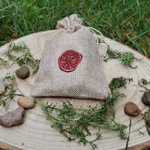 May include: A small burlap pouch with a red wax seal featuring a circular design. The pouch is sitting on a wooden surface with green moss and small stones.