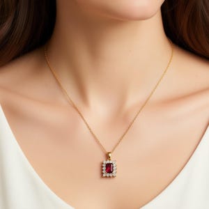 May include: A gold-colored necklace with a rectangular, deep red pendant. The pendant is surrounded by small, clear gemstones. The necklace is worn against a white top.