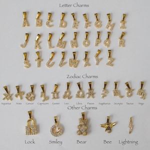 May include: A collection of gold-colored charms, including letters, zodiac signs, and other designs. The charms are adorned with small, sparkling stones. The image includes the words "Letter Charms", "Zodiac Charms", and "Other Charms".