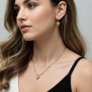 A woman with long, wavy hair wearing a gold necklace and earrings, looking to the side with a thoughtful expression.