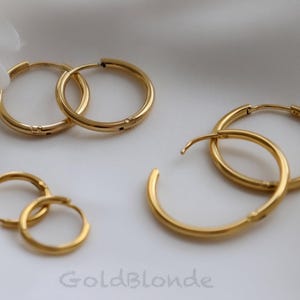 May include: Several pairs of gold-colored hoop earrings of varying sizes are displayed on a white surface. The earrings have a shiny, polished finish. The brand name "Gold Blonde" is visible at the bottom.