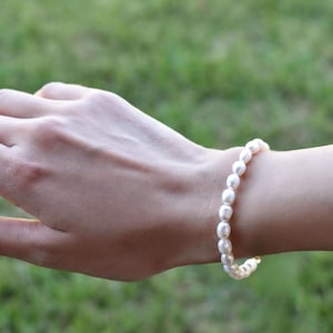 A close-up view of a person's hand wearing a pearl bracelet and a gold-colored bracelet against a blurred green background.