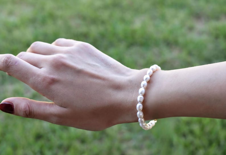 A close-up view of a person's hand wearing a pearl bracelet and a gold-colored bracelet against a blurred green background.