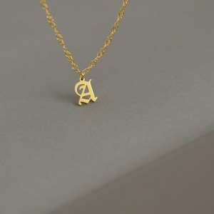 May include: A gold-colored necklace with a pendant in the shape of the letter "A". The pendant is in an elegant, stylized font. The chain is a delicate, twisted rope design. The background is a neutral gray.