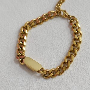 A gold-colored bracelet with a rectangular charm attached to it, resting on a white surface.
