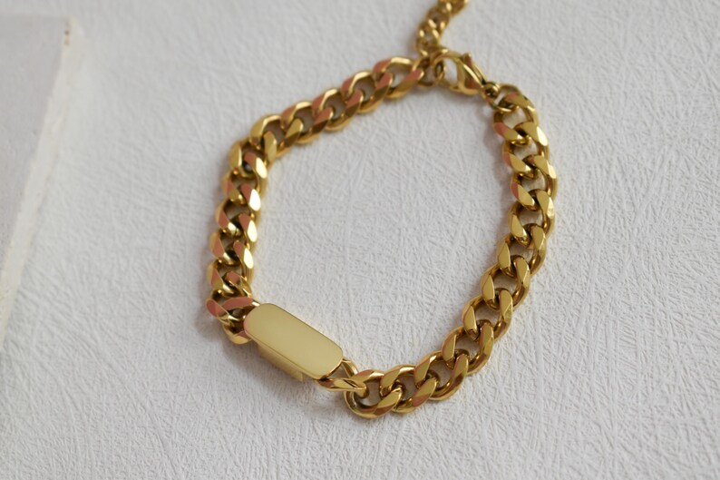 A gold-colored bracelet with a rectangular charm attached to it, resting on a white surface.