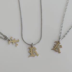 May include: Three silver-toned necklaces featuring pendants shaped like the letter K in an old English font. Each necklace has a different chain style, including a twisted rope chain, a smooth snake chain, and a standard link chain design.
