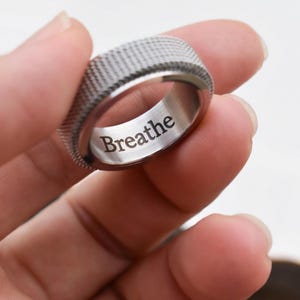 A hand holding a silver ring with the word "Breathe" engraved on it.