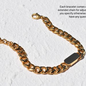 A gold-colored bracelet with a black rectangular charm is shown against a white background.