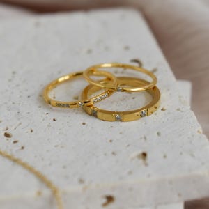 May include: A collection of gold rings with small, clear gemstones. The rings are stacked on a textured, off-white surface. The rings vary in design, with some featuring a band of gemstones and others a simple, smooth band.