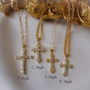 May include: Four gold-colored cross necklaces with clear gemstone accents. Each cross design is unique, with varying shapes and sizes of gemstones. The necklaces hang against a white fabric background, with the letters "F Style", "C Style", "A Style", and "E Style" visible.