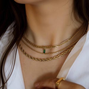 May include: A layered gold-tone necklace set consisting of three distinct chains. The top chain is a flat braided style, the middle chain is a thin rope style with a small rectangular green pendant, and the bottom chain is a thicker rope style.