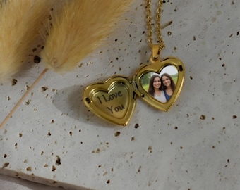 May include: Gold heart-shaped locket necklace, open to reveal a photo of two women. The locket is engraved with "I Love You". The necklace is on a textured surface with dried flowers.