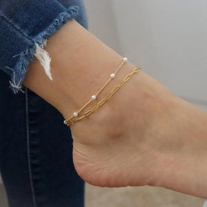 May include: A double-layered gold anklet featuring one delicate chain adorned with small white pearls and a second chain composed of elongated oval links. The jewelry is worn around the ankle, resting above blue denim jeans with a frayed hemline.