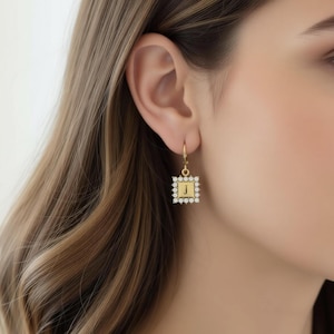 May include: A gold-colored square earring with a small letter "J" in the center, surrounded by a border of small, clear gemstones. The earring is hanging from a gold-colored hook.