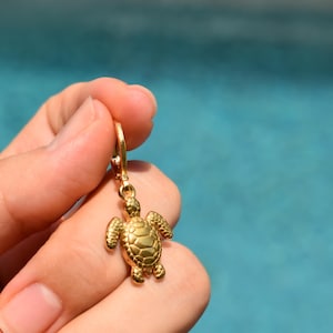 May include: A gold-colored turtle charm earring. The turtle has a detailed shell and flippers. The earring is held up against a blue background.