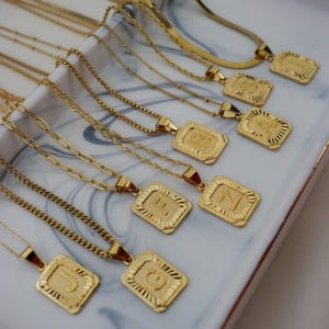 May include: A collection of gold-colored necklaces with rectangular pendants, each featuring a different letter of the alphabet. The necklaces are displayed on a white and gray marbled tray, showcasing various chain styles.