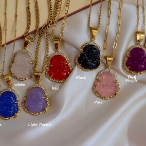 A collection of colorful Buddha pendant necklaces with various gemstones and gemstone settings, displayed on a white fabric background.