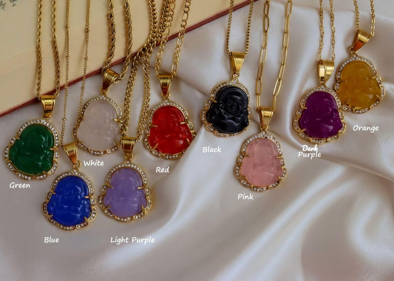 A collection of colorful Buddha pendant necklaces with various gemstones and gemstone settings, displayed on a white fabric background.