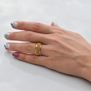 A close-up view of a hand with a gold ring and a marble nail polish design, resting on a white surface.