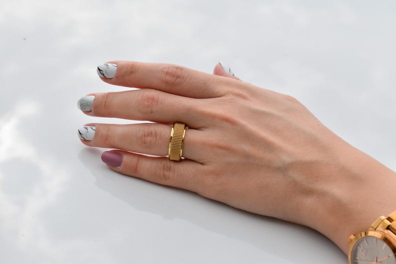 A close-up view of a hand with a gold ring and a marble nail polish design, resting on a white surface.