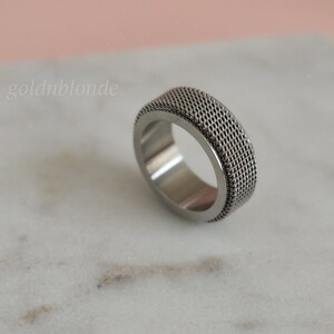 A silver ring with a textured surface sits on a white marble surface.
