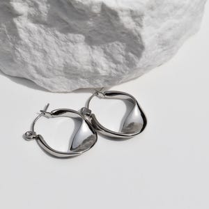 May include: A pair of silver-toned hoop earrings with a unique, twisted design. The earrings are displayed on a white surface with a large, textured white stone in the background. The earrings have a polished, reflective surface.