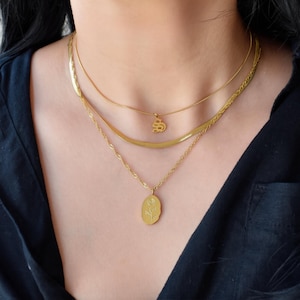 May include: A layered gold-tone necklace set consisting of three distinct chains. The top chain is a thin snake chain with a small letter pendant, the middle is a flat herringbone chain, and the bottom is a twisted rope chain with an oval pendant.