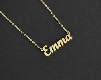 May include: A gold-colored necklace featuring a delicate chain and a nameplate pendant in a cursive script font that spells out the name Emma. The metal has a polished finish and the pendant is attached to the chain on both sides.