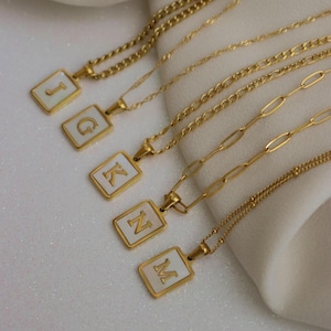 May include: Gold-toned necklaces with rectangular pendants featuring white backgrounds and gold lettering of the letters J, G, K, N, and M. The chains have various designs, including curb and link styles.
