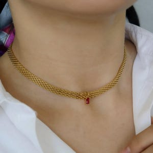 May include: A gold-toned mesh chain choker necklace featuring a single, small, rectangular red gemstone pendant suspended from the center. The necklace has a woven, textured metal link design that sits closely around the base of the neck.