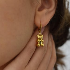 May include: Gold-colored teddy bear charm earrings. The earrings feature a small, detailed teddy bear pendant hanging from a hoop. The bear has a rounded body and head with small arms and legs. The earrings are worn on the ear.