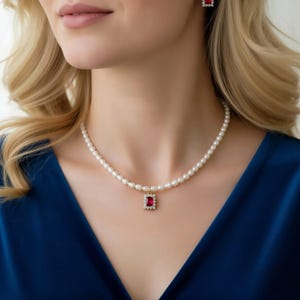 May include: A necklace featuring a strand of small white pearls with a central rectangular pendant. The pendant contains a deep red gemstone surrounded by a border of small clear stones, all set in a gold-toned metal frame.