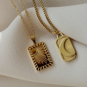 May include: Two gold-colored pendant necklaces. One rectangular pendant features a sunburst design, and the other has a crescent moon and sunburst. Both necklaces are on delicate chains, set against a white background.