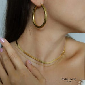 A close-up of a woman's neck and chest area, featuring a gold necklace, large hoop earrings, and a ring on her finger.