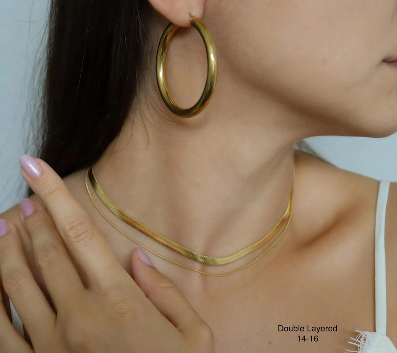 A close-up of a woman's neck and chest area, featuring a gold necklace, large hoop earrings, and a ring on her finger.