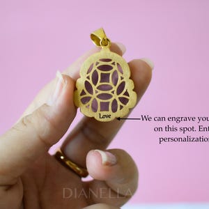 A gold pendant with a floral design is being held in a hand against a pink background.