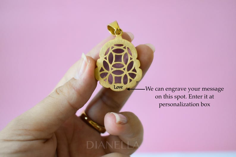 A gold pendant with a floral design is being held in a hand against a pink background.
