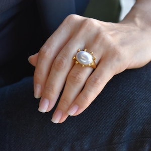 May include: A gold ring featuring a large, oval, white pearl. The ring has a decorative gold band with small, round accents. The ring is on a hand, with a dark background.