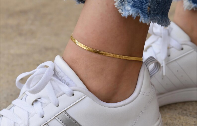 A close-up view of a person's ankle wearing a gold snake chain bracelet and white sneakers.