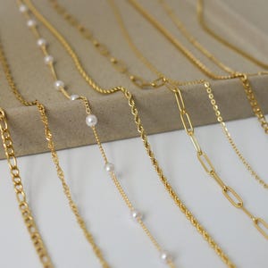 May include: A collection of gold-colored necklaces displayed on a neutral-toned tray. The necklaces feature various chain styles, including delicate chains with small pearls, rope chains, and link chains. The background is a soft, neutral color.