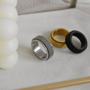 three rings of different colors and designs, including a silver ring, a gold ring, and a black ring, placed on a white surface next to a white candle.