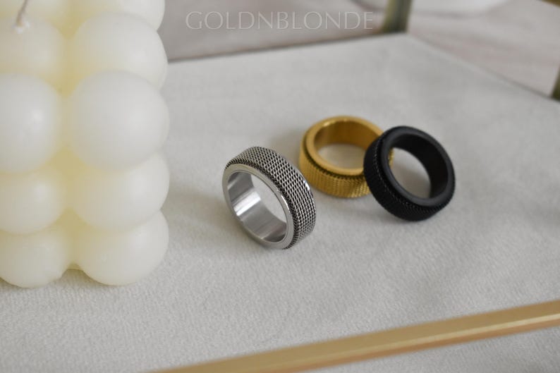 three rings of different colors and designs, including a silver ring, a gold ring, and a black ring, placed on a white surface next to a white candle.