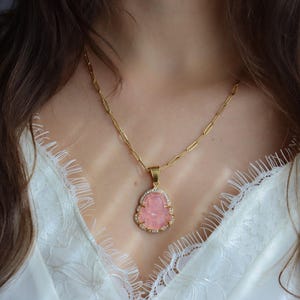 A close-up of a woman's neck and chest wearing a gold necklace with a pink gemstone pendant.