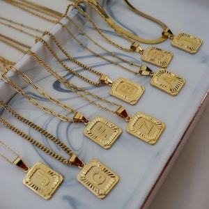 May include: A collection of gold-toned necklaces featuring rectangular pendants with embossed capital letters. Each pendant has a textured border and is suspended from various chain styles, including cable, curb, and herringbone link designs.